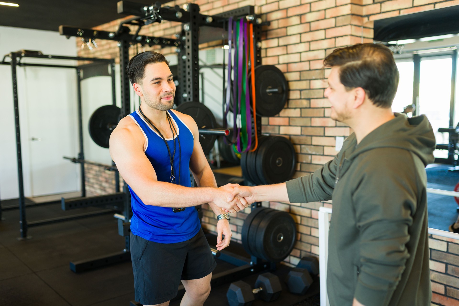 Muscular fitness trainer greeting client with handshake, gym equipment background, embodying workout collaboration and motivation